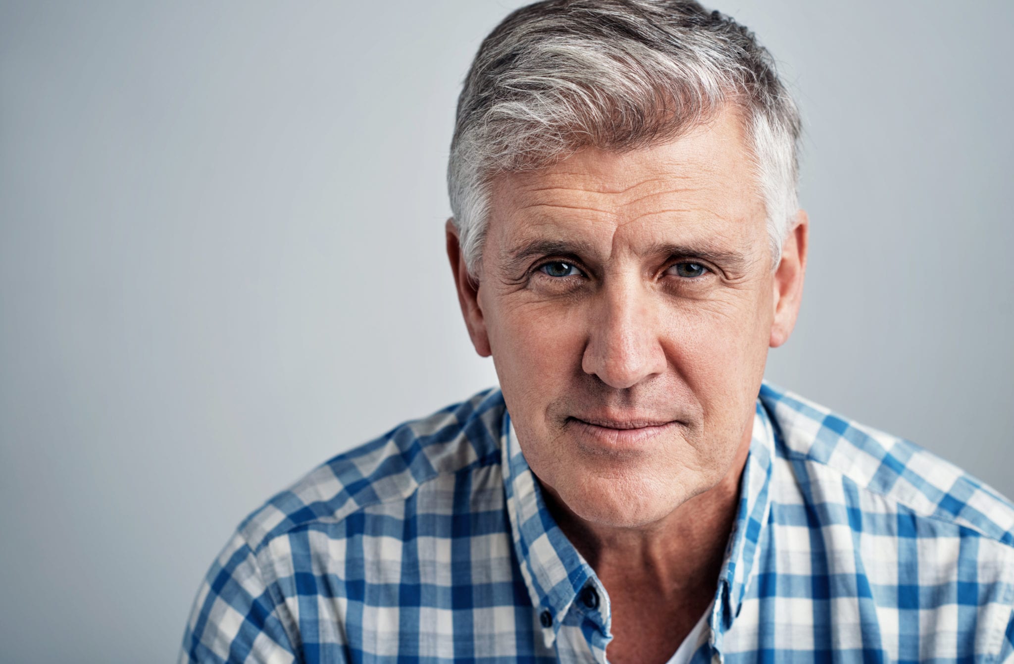 headshot of seated man in plaid shirt with grey hair with a slight smirk on his face, against grey background