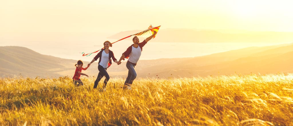 family running together on high grass field with skies and more hills in the background as father holds a kite