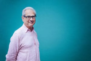 An image of a happy elderly man wearing a pink button down shirt and glasses, with a dark teal background.