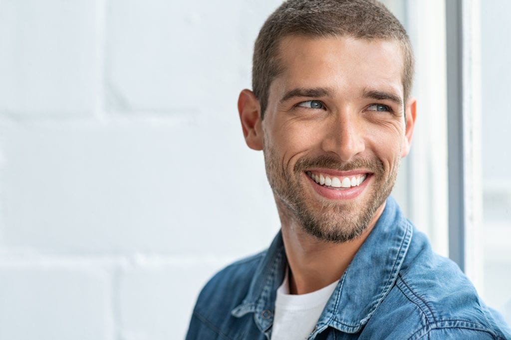 Headshot of well groomed man in denim jacket turning towards camera and smiling widely with blurred background showing a window and grey wall